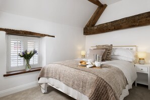 A bedroom with a bed, bedside tables, a window with blinds, and a vase of flowers. - The Mews Hideaway -  a cottage that sleeps 4 guests  in 2 bedrooms (Fairford)