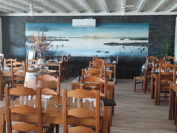 A restaurant with wooden tables and chairs, a large painting of a landscape on the wall, and an air conditioning unit.