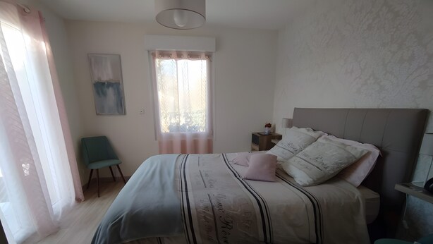 Een slaapkamer met een bed, een stoel, een nachtkastje, een raam met gordijnen en een wandlamp.