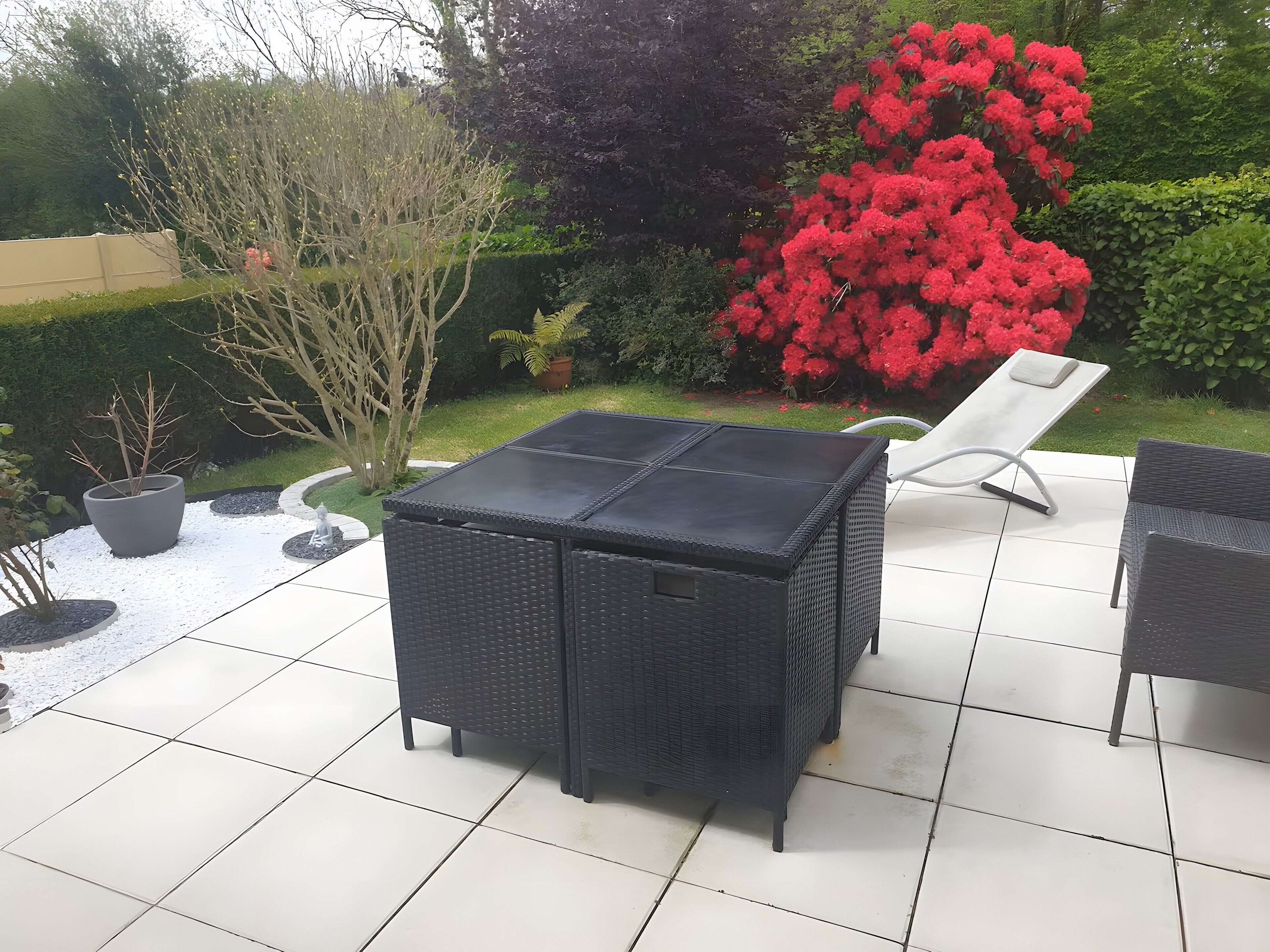 Eine Terrasse mit einem schwarzen Korbstuhl-Esstisch und -Stühlen, umgeben von einem Garten mit verschiedenen Pflanzen und einem roten Busch mit Blüten.