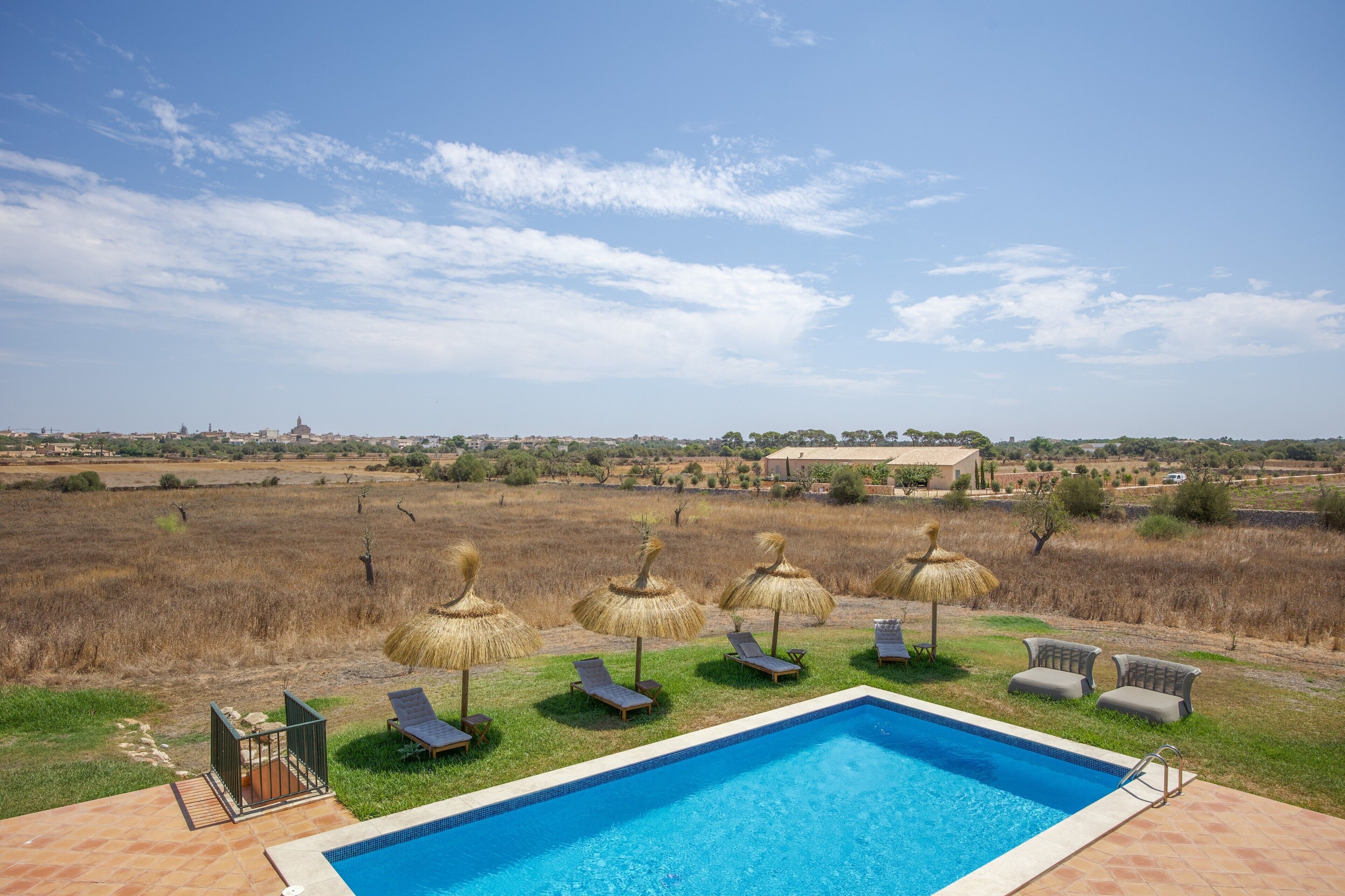 A swimming pool with lounge chairs and thatched umbrellas, surrounded by a grassy area with a view of a distant town.