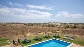 A swimming pool with lounge chairs and thatched umbrellas, surrounded by a grassy area with a view of a distant town.