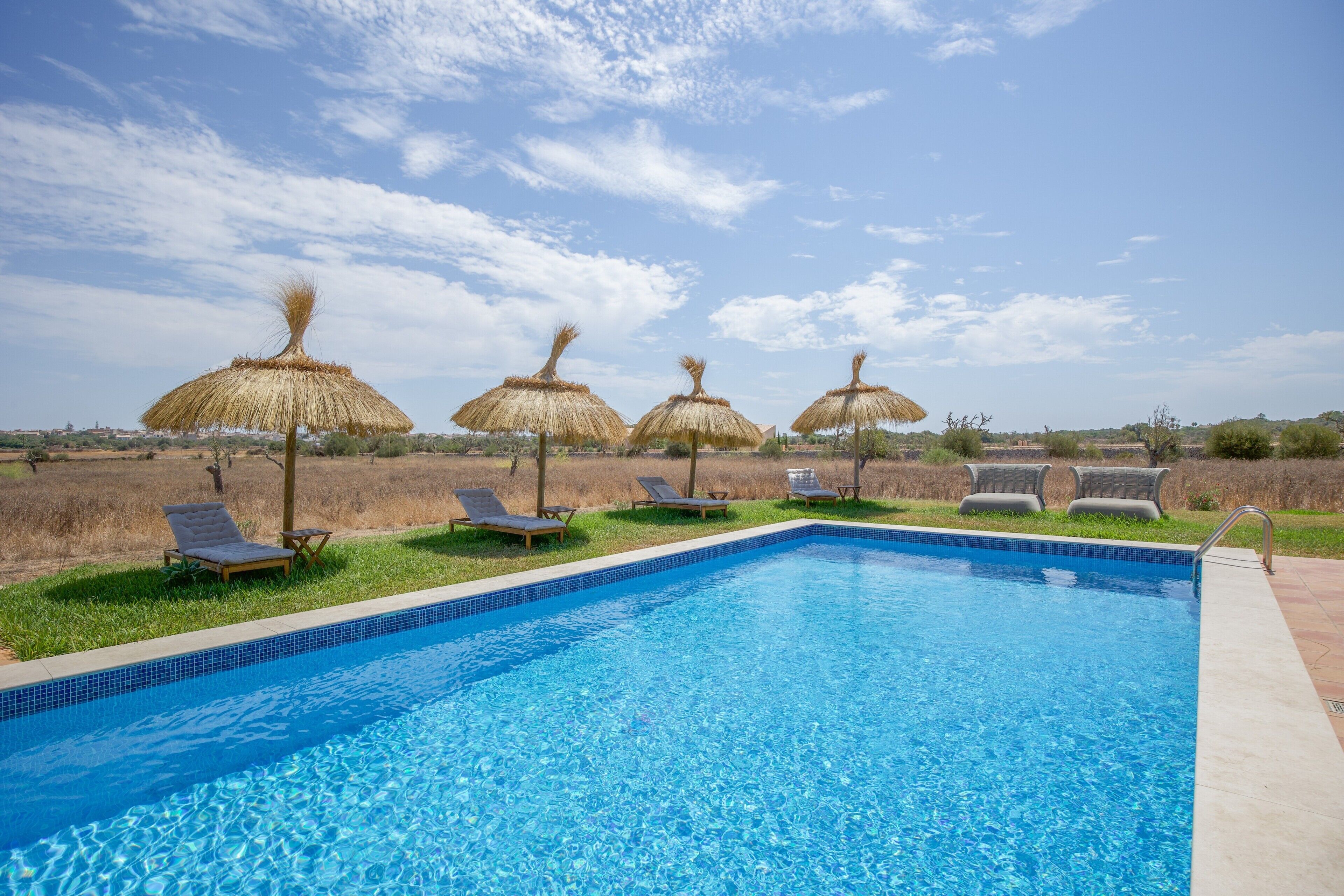 Une piscine entourée de chaises longues et de parasols en chaume, le tout bordé d’une pelouse et surplombé par un ciel dégagé.