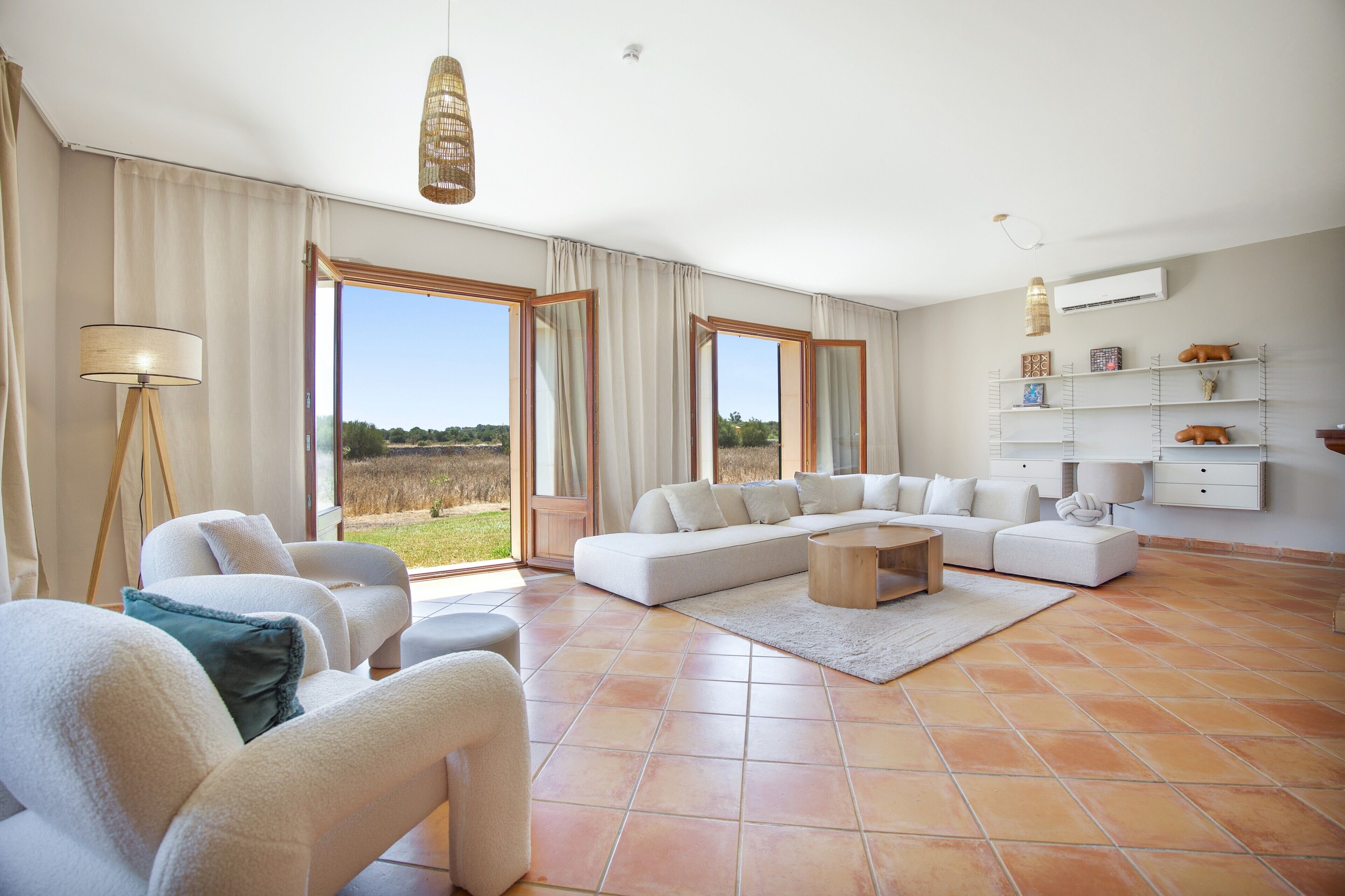 A spacious living room with a large sectional sofa, a coffee table, and a floor lamp. There are large windows offering a view of the outdoors.