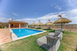 Una piscina con sillones de descanso y sombrillas, un pequeño edificio con techo de tejas y una zona ajardinada.
