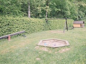 A grassy outdoor area with a wooden swing set, a small wooden structure, and a hexagonal sandbox.