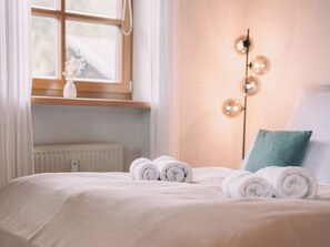 A neatly made bed with rolled towels, a bedside lamp, and a window with curtains.