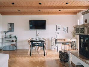 A modern living room with a wooden ceiling, a flat-screen TV mounted on the wall, a dining table with chairs, a bookshelf, and a fireplace.