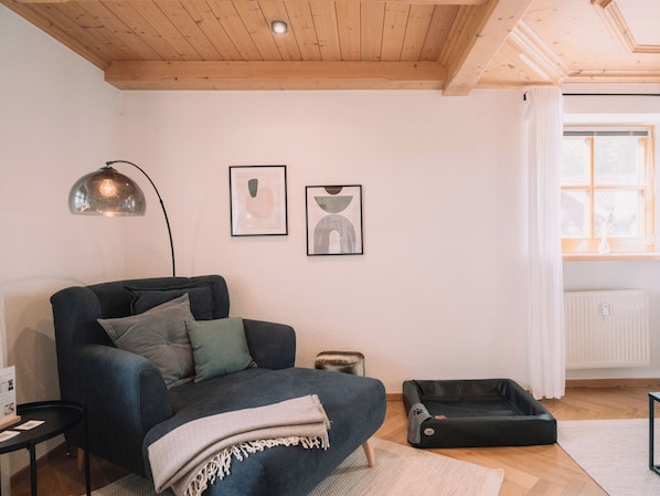 A cozy living room with a dark sofa, a pet bed, a lamp, and two framed artworks on the wall.