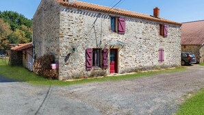 Una casa de piedra con puerta roja, dos ventanas con contraventanas y un camino de entrada de grava.