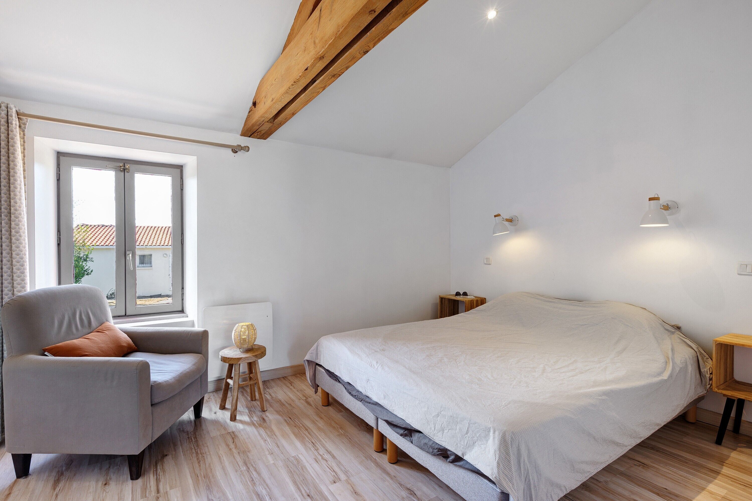 Ein Schlafzimmer mit einem Bett, einem Sessel, einem kleinen Tisch, einem Fenster und einer Decke mit freiliegenden Balken.