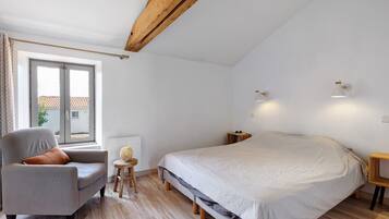 A bedroom with a bed, a chair, a small table, a window, and a ceiling with exposed beams.