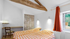 Un dormitorio con una cama, una silla, un escritorio, una ventana con cortinas y una viga de techo de madera.
