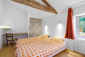 Un dormitorio con una cama, una silla, un escritorio, una ventana con cortinas y una viga de techo de madera.