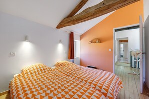 A bedroom with a bed, two bedside lamps, a window with curtains, and a wooden ceiling beam.