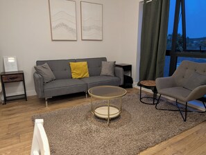 Ein modernes Wohnzimmer mit einem grauen Sofa, einem runden Couchtisch, einem grauen Sessel und einem großen Fenster mit Vorhängen.