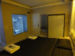 A hotel room with a bed, a window with a view of buildings, a door, and a small table with a lamp. - ÇANKAYA SUIT (Çankaya)