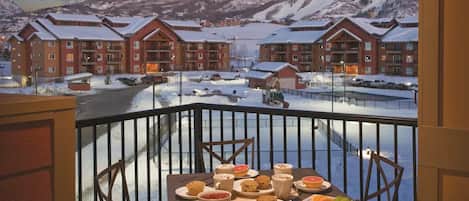 A balcony with a table set for breakfast, overlooking a snow-covered landscape with buildings.