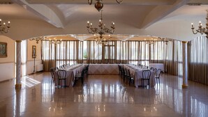 Une vaste salle de banquet, avec une longue table dressée pour un événement officiel, des lustres somptueux et de grandes fenêtres ornées de rideaux.