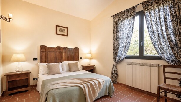 Een slaapkamer met een houten hoofdeinde, twee bedlampen, een bed met een beige deken, een raam met behang en een stoel.