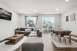 A modern living room with a grey sectional sofa, a wooden coffee table, and a dining area with a wooden table and chairs. - Family Vacation Home - DWTN - Airhockey - Spacious (Vancouver)