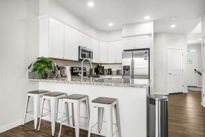 A modern kitchen with white cabinetry, a stainless steel refrigerator, a microwave, and a granite countertop. There are four white bar stools and a potted plant. - Family Vacation Home - DWTN - Airhockey - Spacious (Vancouver)