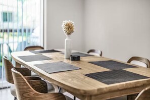 Une table de conférence en bois, sur laquelle reposent des sets de table noirs, un vase blanc contenant des fleurs séchées et un objet noir.