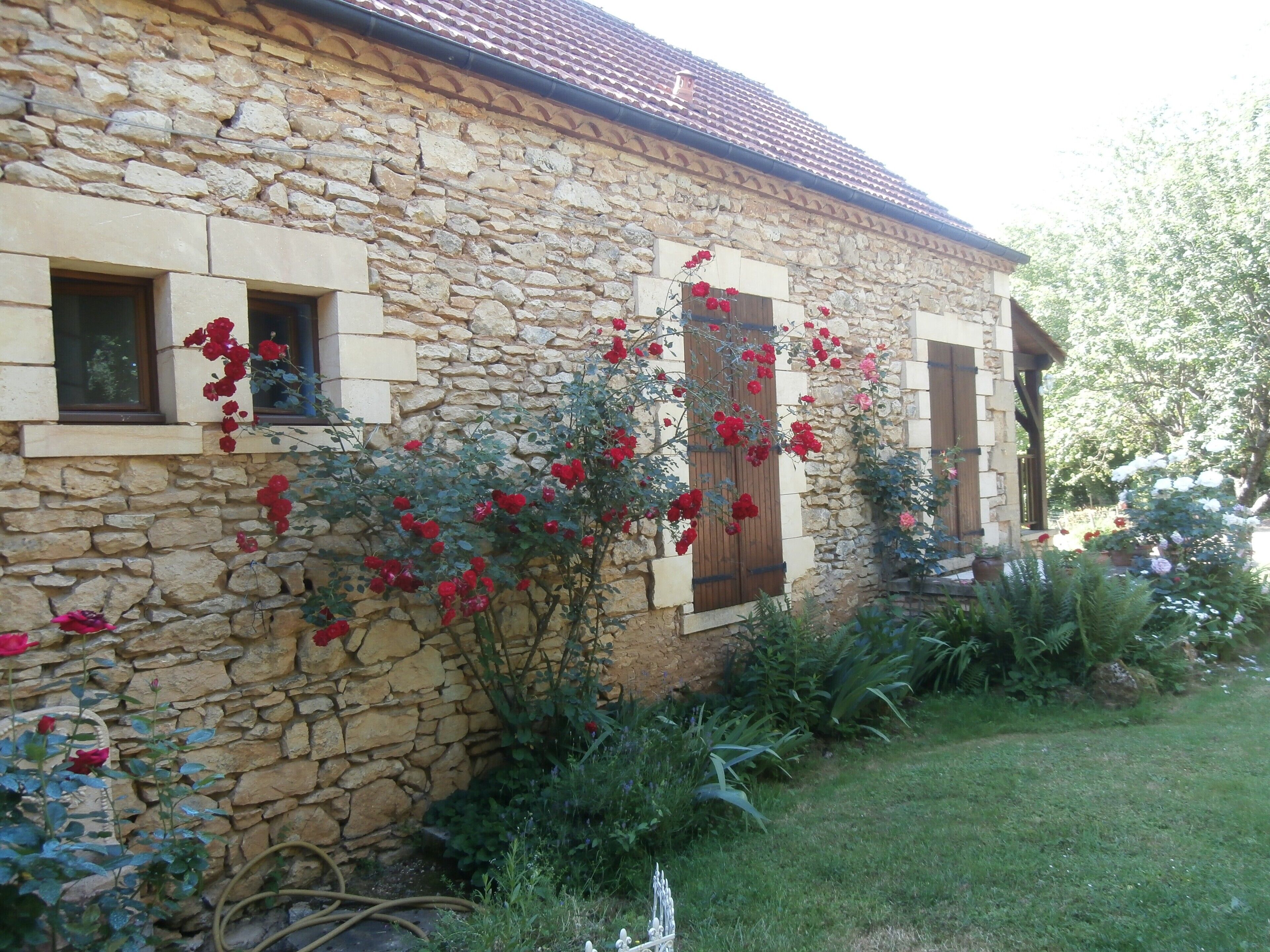 A stone building with a garden, featuring a stone wall, wooden shutters, and blooming red flowers.