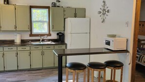 Uma cozinha com armários verde-claros, uma geladeira branca, um micro-ondas e uma janela com vista para árvores. Há três banquetas de madeira na ilha da cozinha.