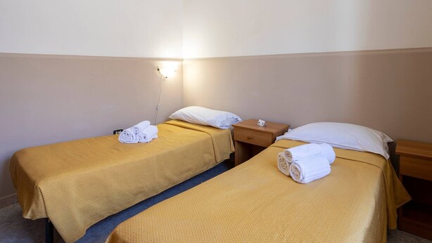 Et hotelværelse med to enkelt senge, hver med en foldet håndklæde og en pude, et natbord og en lampe på væggen.