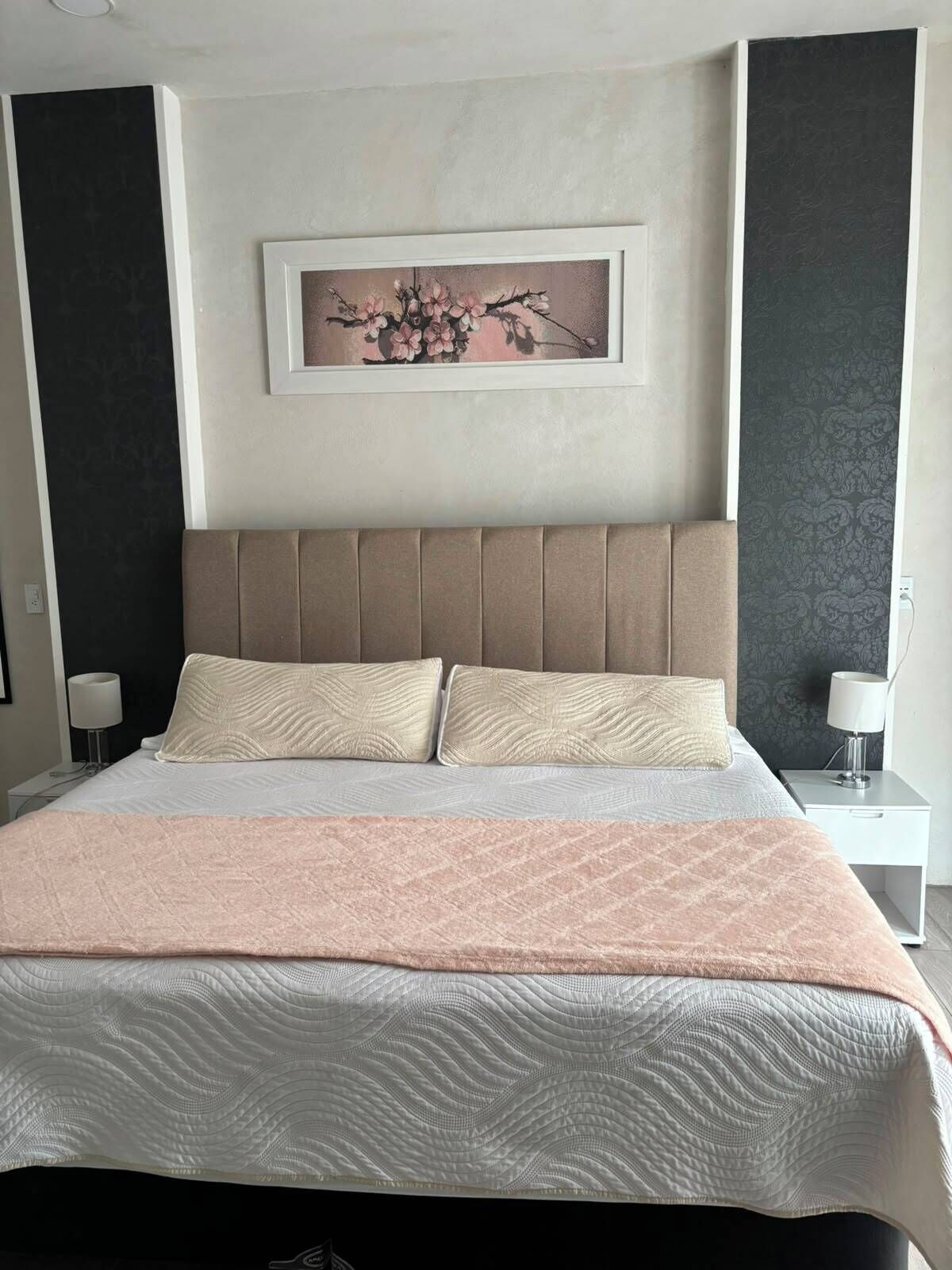 Una cama bien hecha con una colcha estampada y dos almohadas, una obra de arte colgada en la pared encima y una mesita de noche con una lámpara.