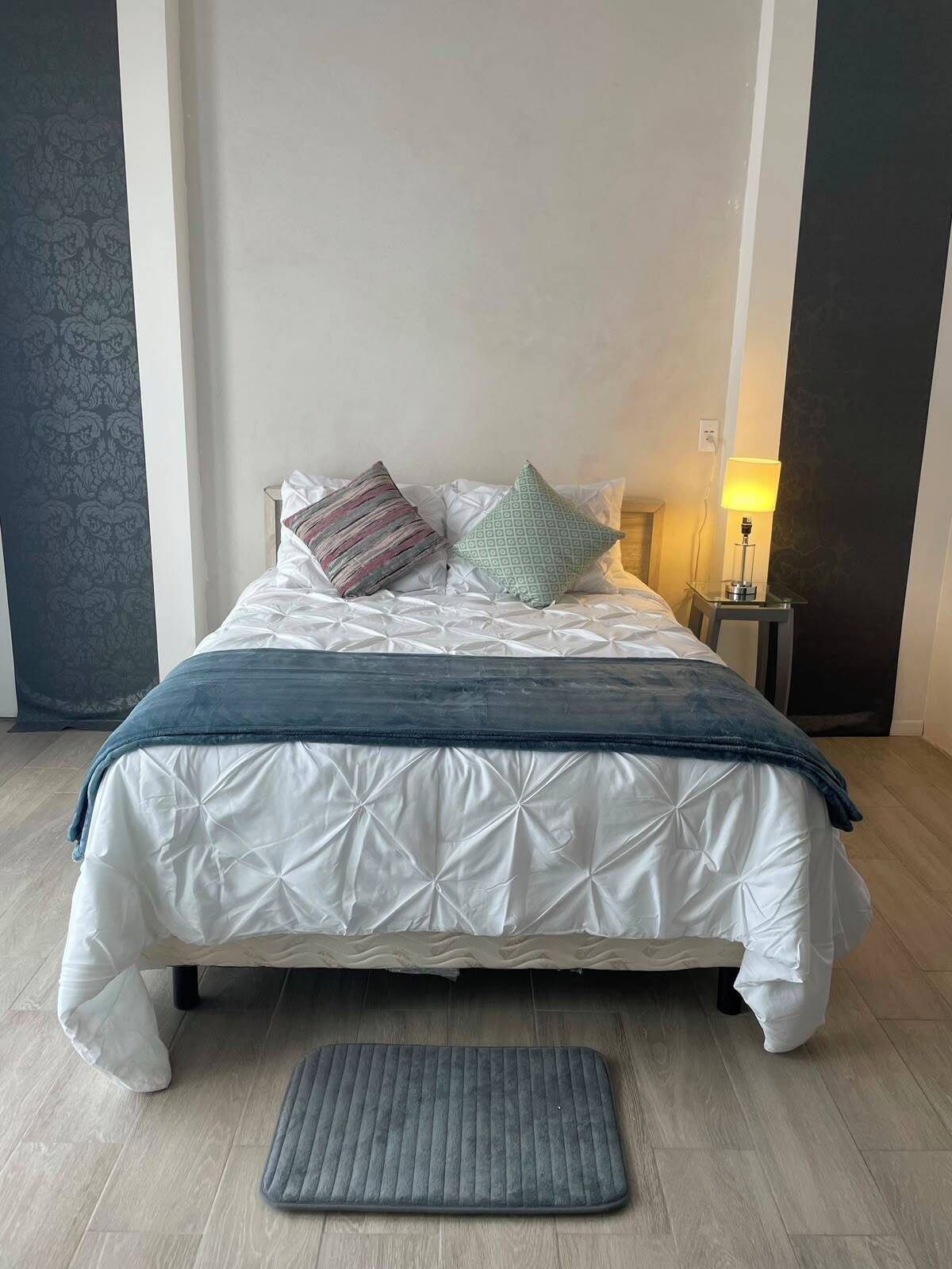 A neatly made bed with a white duvet and a blue throw blanket, accompanied by two pillows, a bedside lamp, and a grey mat on a wooden floor.