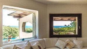 A room with a view of the sea through a window, a sofa with cushions, and a framed picture on the wall. - villa Passione (Palau)