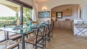 A dining area with a glass table and chairs, a kitchen with wooden cabinets, and a view of greenery outside. - villa Passione (Palau)