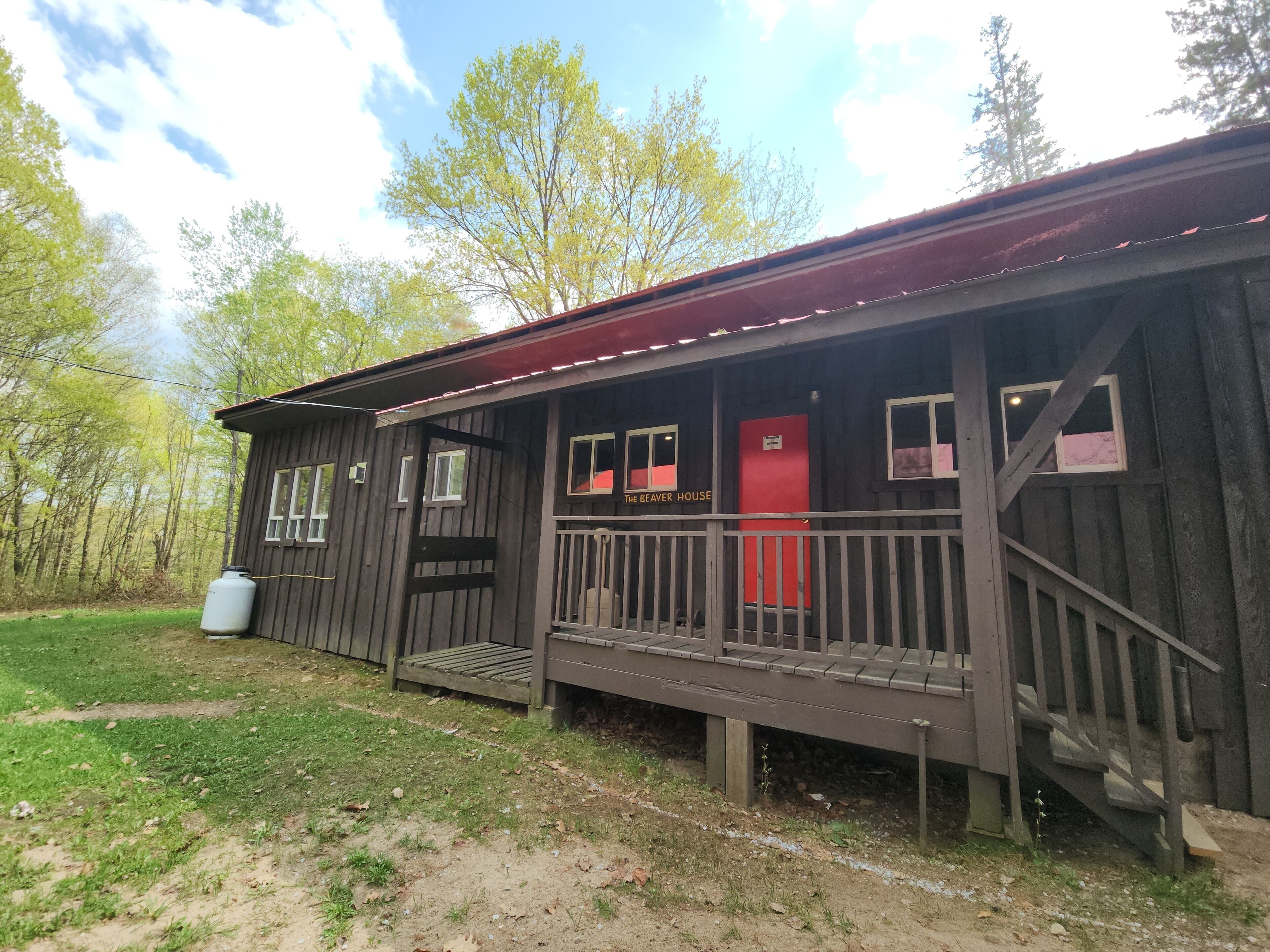 A rustic wooden cabin with a porch, a red door, and a small staircase leading to the entrance.