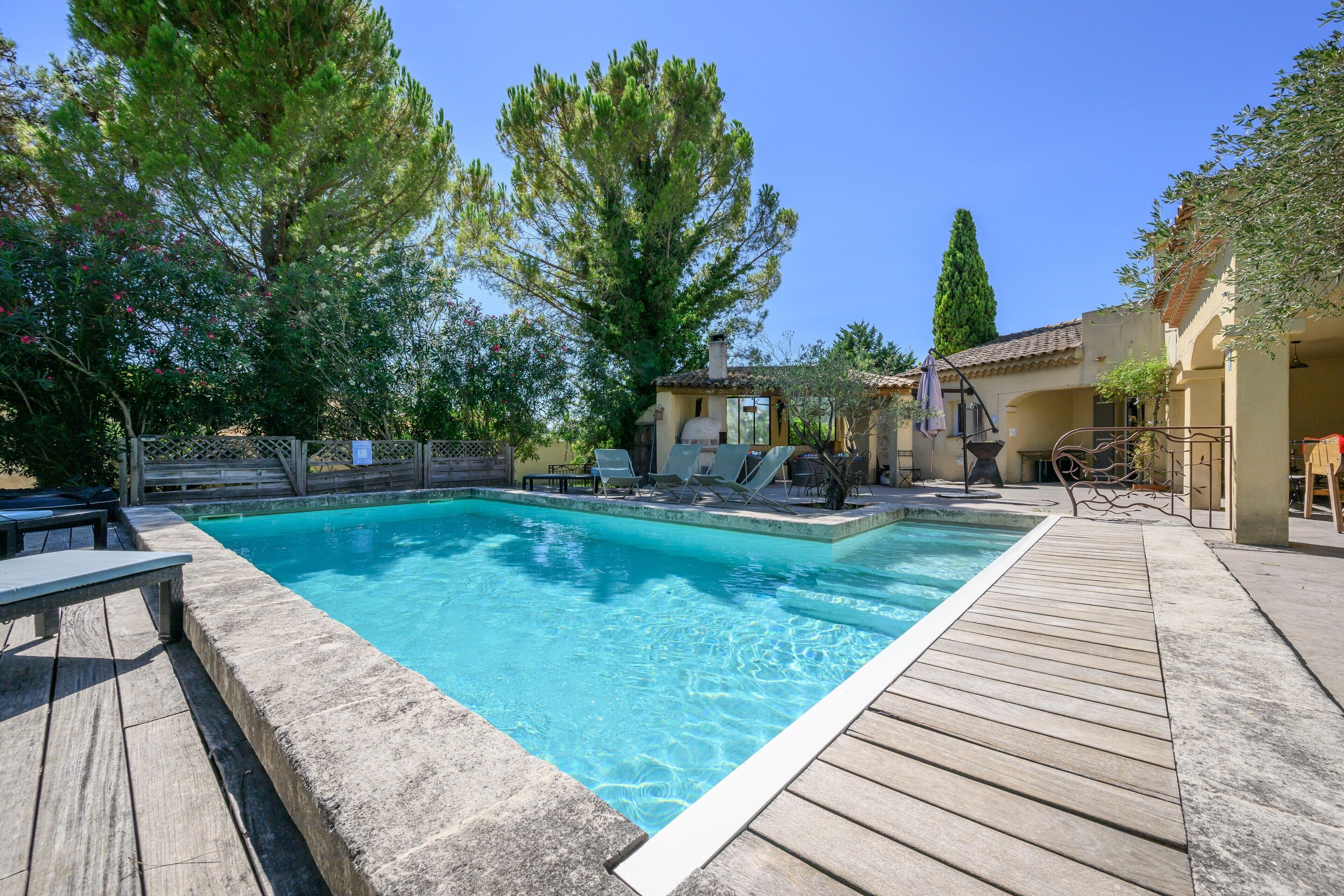 Une piscine rectangulaire entourée d’une terrasse en bois, un petit bâtiment doté d’une cheminée et des arbres en arrière-plan.