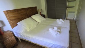 Ein ordentlich bezogenes Bett mit weißen Leinen und zwei aufgerollten Handtüchern, ein Holz-Kopfteil, ein Nachttisch mit Lampe und ein Kleiderschrank im Hintergrund.