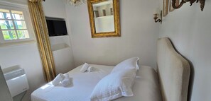 Ein ordentlich bezogenes Bett mit weißen Leinen und Handtüchern, ein spiegel mit goldener Rahmung und ein Fenster mit Vorhängen.