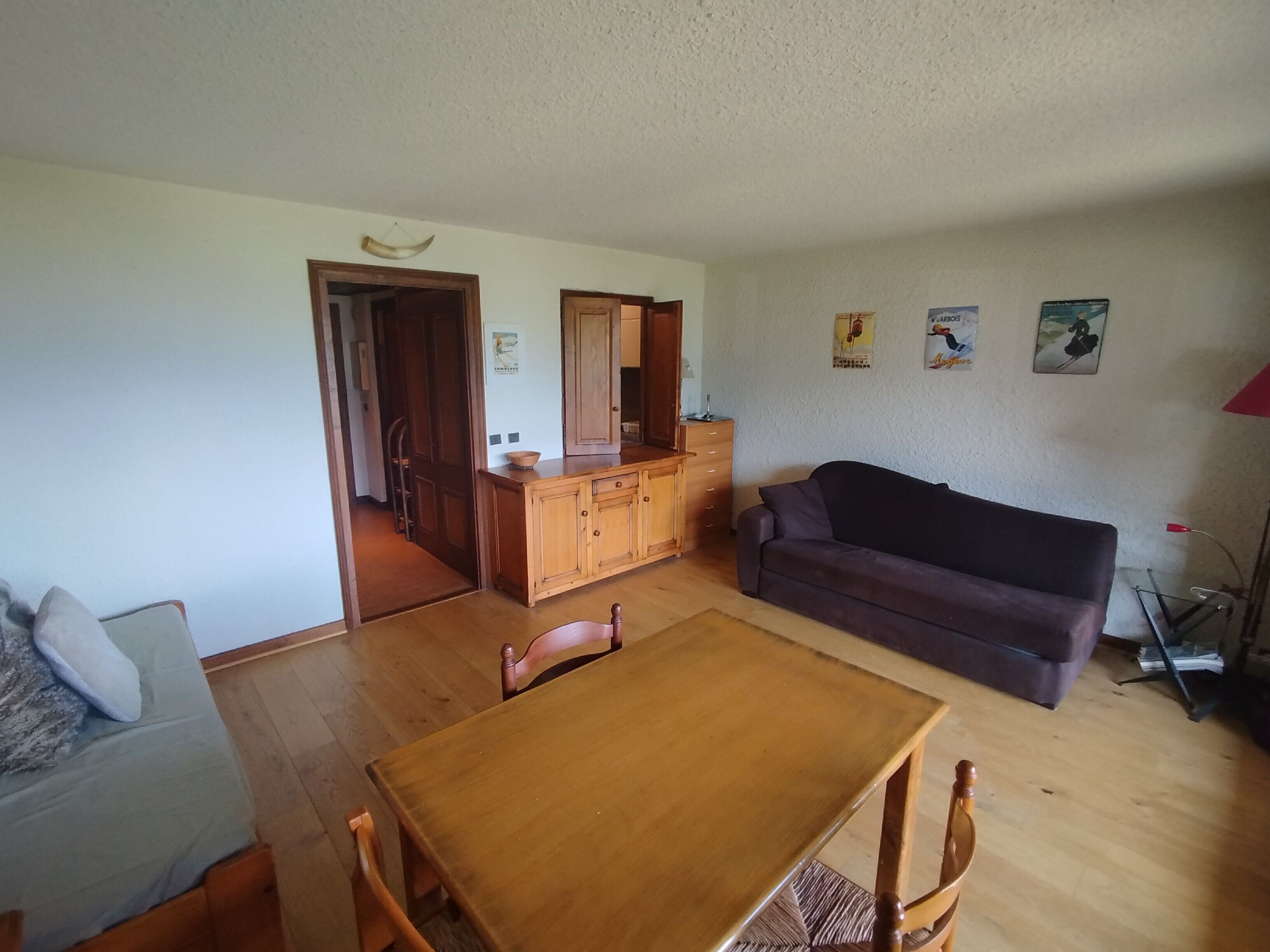 A living room with a wooden dining table, a dark sofa, and a kitchen area with wooden cabinets.