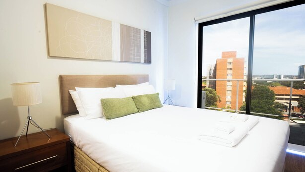 Ein Hotelzimmer mit Bett, Nachttisch, Lampe und einem großen Fenster mit Blick auf die Stadt.