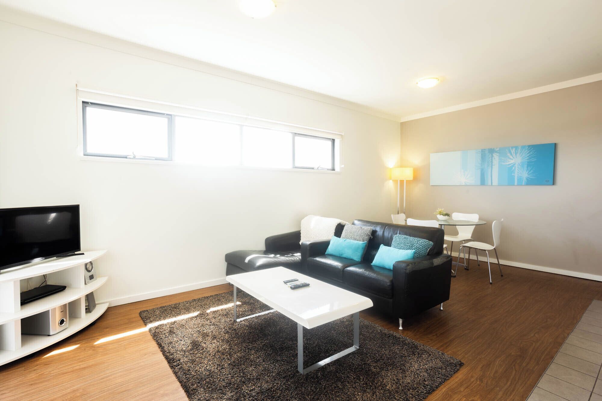 A modern living room with a flat-screen TV, a sofa with cushions, a coffee table, and a dining area with a table and chairs.