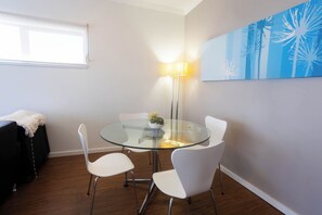 A modern dining area with a glass table, white chairs, a floor lamp, and a blue abstract painting on the wall. - Lovely 2BR Apartment in West Perth with Parking! (West Perth)