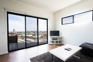 A modern living room with a large sliding glass door leading to an outdoor balcony, a flat-screen TV on a white stand, a white coffee table, and a black sofa. - Lovely 2BR Apartment in West Perth with Parking! (West Perth)
