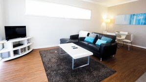 A modern living room with a flat-screen TV, a black leather sofa with blue cushions, a white coffee table, and a dining area with a small table and chairs.