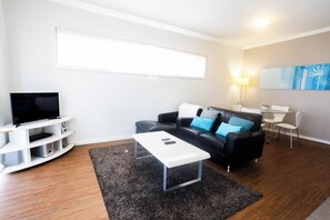 A modern living room with a flat-screen TV, a black leather sofa with blue cushions, a white coffee table, and a dining area with a small table and chairs. - Lovely 2BR Apartment in West Perth with Parking! (West Perth)