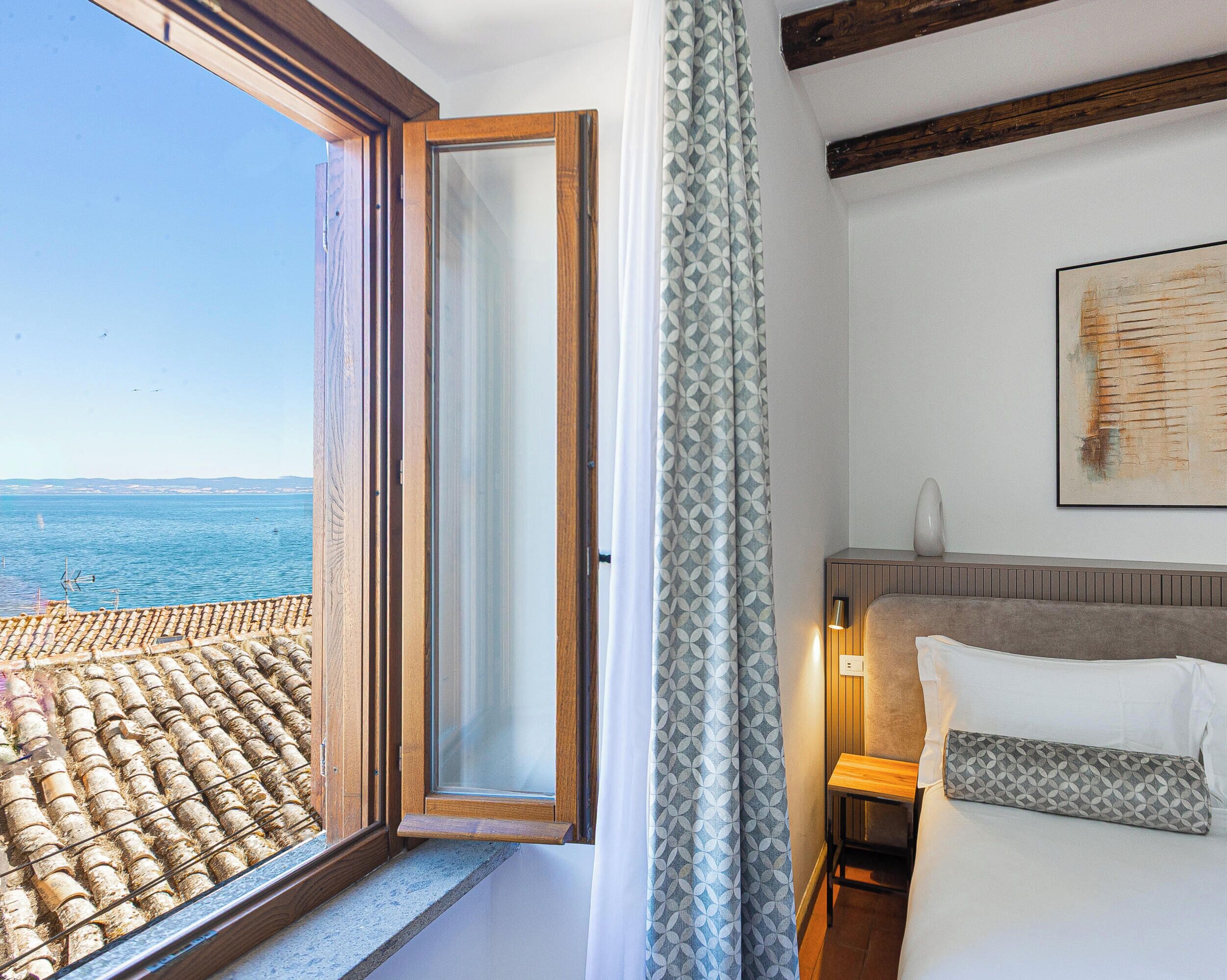 Habitación de hotel con un ventanal grande, una cama con ropa blanca, una mesita de noche con lámpara y un cuadro en la pared.