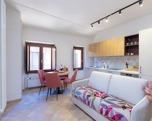 A modern kitchen with a built-in oven, a dining area with a table and chairs, and a sofa with a colorful cushion. - Marta Lake Albergo Diffuso & Spa (Marta)