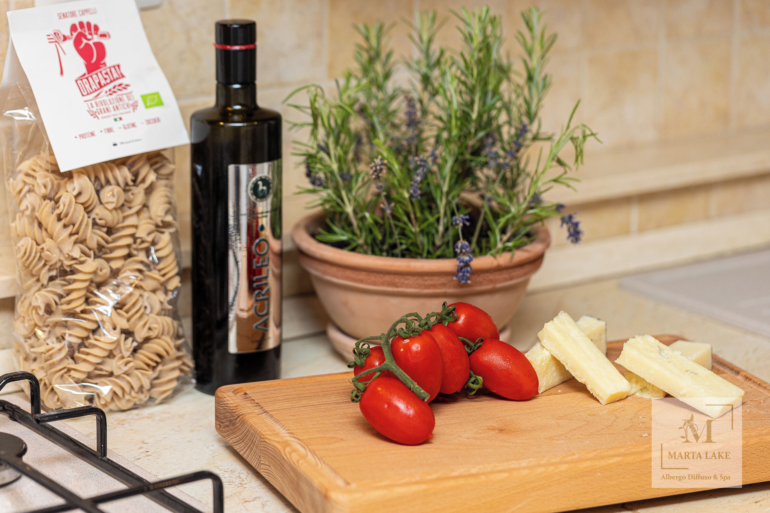 A kitchen counter with a cutting board, tomatoes, cheese, a bottle of olive oil, a jar of pasta, and a potted herb plant.