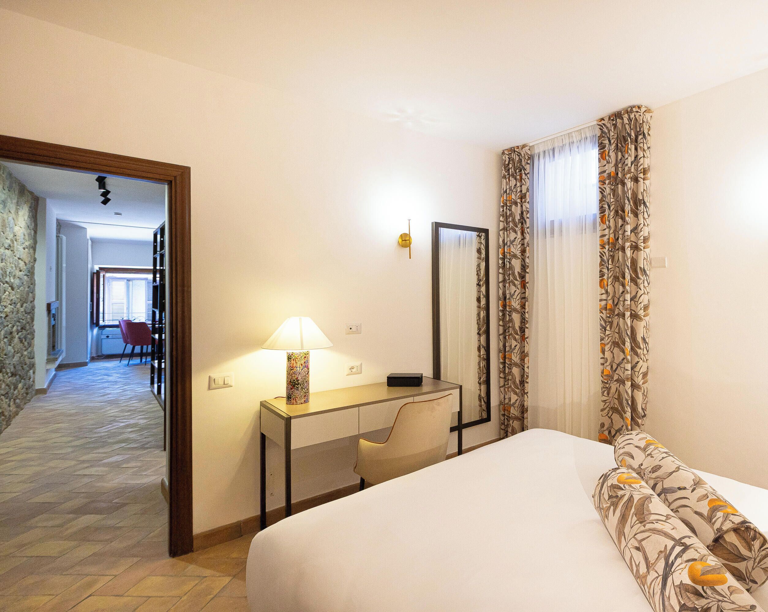 A hotel room with a bed, desk, chair, and a mirror. There is a door leading to another room.