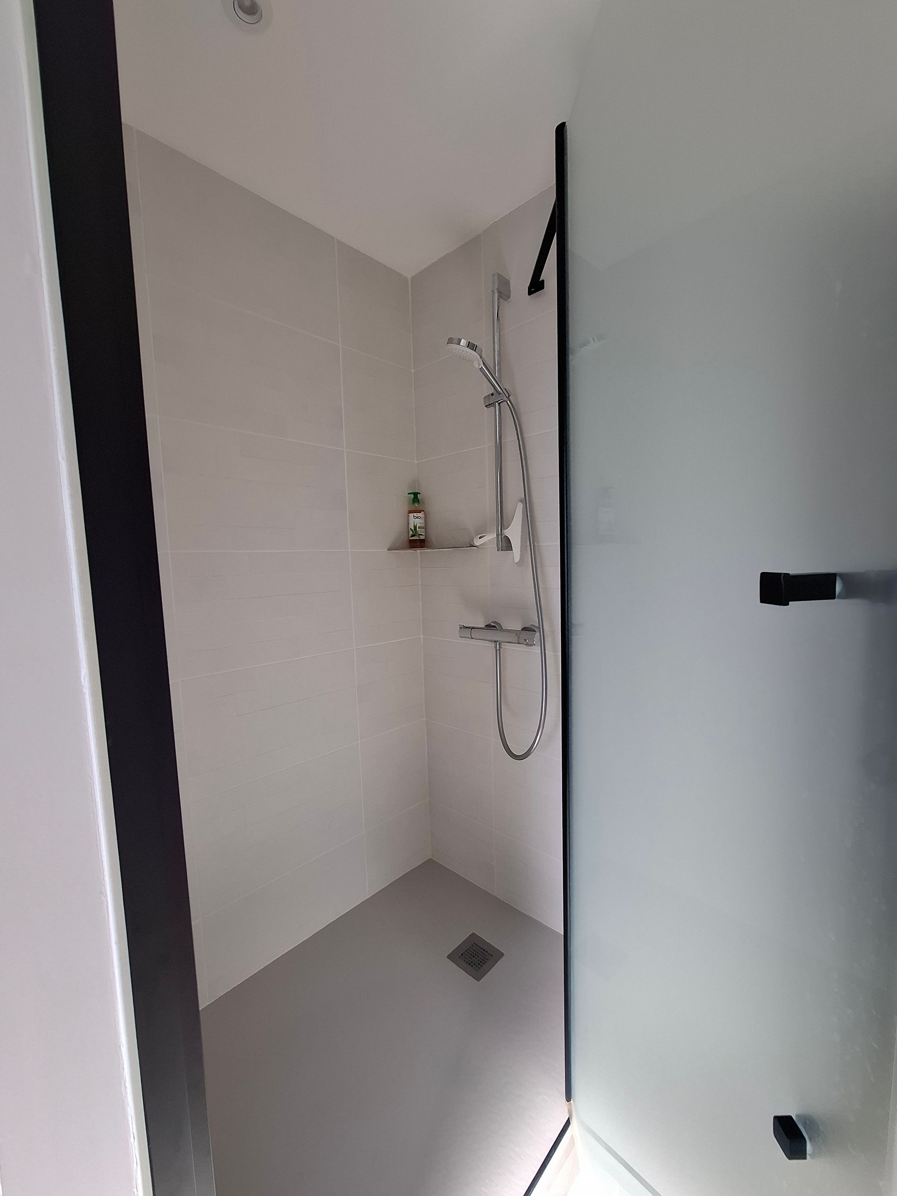 A modern bathroom with white tiles, a shower with a handheld showerhead, and a small shelf with a bottle.